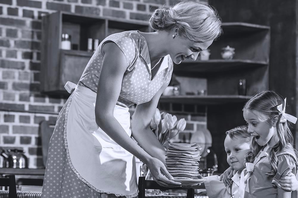 A woman holding a plate of pancakes in front of two children that look excited